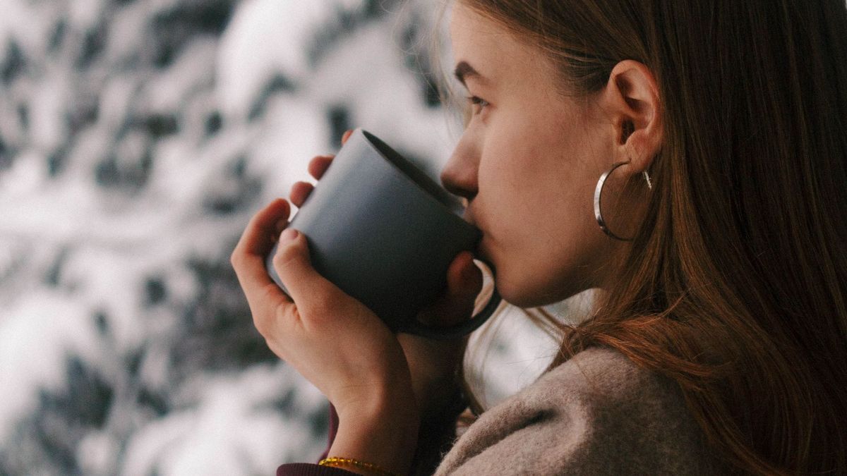 A woman in a warm sweater sips a hot drink beside a window with snowy trees outside